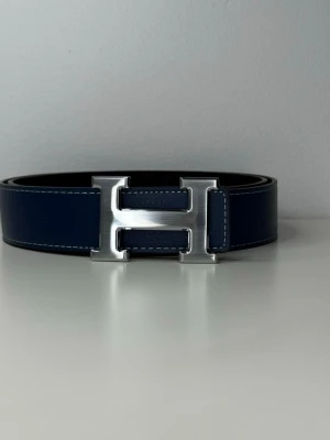  mörkblått skärp med H-spänne - Stilrent mörkblått skärp med ikoniskt H-spänne i silverfärgad metall. Skärpet är tillverkat i exklusivt skinn och har synliga vita sömmar längs kanterna. Perfekt accessoar för att lyfta din outfit.