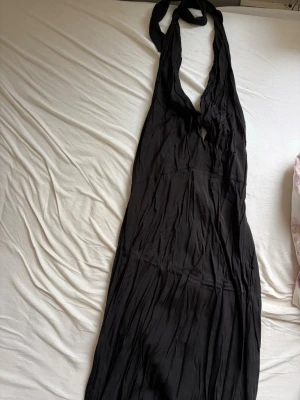 Halterneck satin Svart långklänning med knyt - Svart långklänning med djup v-ringning och knyt i nacken. Klänningen har en mjuk, flytande passform och är ärmlös. Perfekt för sommarens alla tillfällen och ger en elegant siluett.