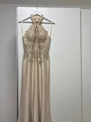 Champagnefärgad långklänning med spets - Elegant långklänning i champagnefärg med halterneck och detaljerad spets upptill. Klänningen har en figurnära överdel med broderade mönster och en slät, fallande kjol i satinliknande material. Perfekt för bal eller festligare tillfällen.                                                           Klänningen är använd 1 gång. Förvarad bra. Hör av dig för fler bilder (bilder med kläning på)                   Original pris: 4300kr