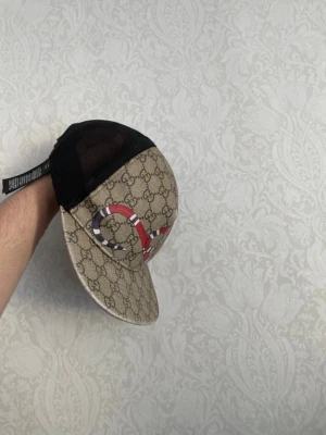 Gucci keps med GG-mönster och orm - Säljer en beige och svart keps från Gucci med klassiskt GG-mönster och en röd, vit och blå orm broderad på framsidan. Kepsen har mesh på baksidan och justerbar rem. Perfekt för dig som vill sticka ut med en lyxig accessoar.