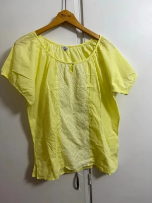 Gul kortärmad blus från Kaffe - Ljusgul blus från Kaffe i modellen Perla Blouse. Tillverkad i 100% bomull med korta ärmar och rundad halsringning. Blusen har en dekorativ panel framtill och avslappnad passform, perfekt för en fräsch och somrig look.