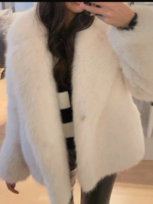 Vit fluffig fuskpälsjacka - Säljer en supermjuk och fluffig vit fuskpälsjacka med bred krage och avslappnad passform. Jackan har långa ärmar och stängs framtill. Perfekt för dig som vill ha en trendig och varm look under vintern.