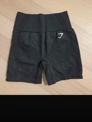  träningsshorts Gymshark svart/grå Size: S - Säljer ett par svarta högmidjade träningsshorts från Gymshark. De har en tight passform och är tillverkade i ett syntetmaterial som är perfekt för träning. Diskret logga på sidan och snygga detaljer längs benen. Perfekt för gymmet eller löpning. Aldrig använt