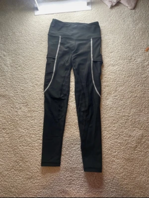 Maya delorez ridbyxor! - Säljer ett par svarta ridbyxor med hög midja och praktiska sidofickor. Tightsen har vita kontrastsömmar framtill och mönstrade detaljer baktill. Perfekta för ridning, med stretchigt material. Super smidiga för ridning!! 4x junior