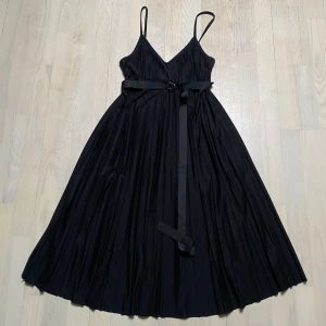 Svart knä lång klänning med bälte - Elegant svart klänning med smala axelband och v-ringning. Klänningen har ett stilrent bälte i midjan och ett plisserat tyg som ger en fin rörelse. Perfekt för en kväll ute eller en speciell tillställning. Tommy Hilfiger strl S