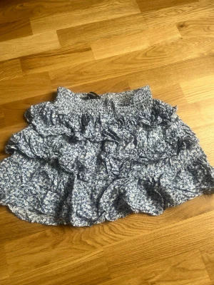Blommig volangkjol från Sister - Supersöt kort kjol från Sister i blått och vitt med blommigt mönster. Kjolen har flera volanger och en smockad midja som ger en bekväm och snygg passform. Perfekt för dig som gillar en feminin och trendig stil.