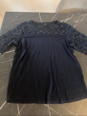 Marinblå topp med spetsdetaljer - Snygg marinblå topp med spets över axlar och ärmar. Toppen har rund halsringning och korta ärmar i genombruten spets som ger en elegant touch. Perfekt att styla med jeans eller kjol för en trendig look.