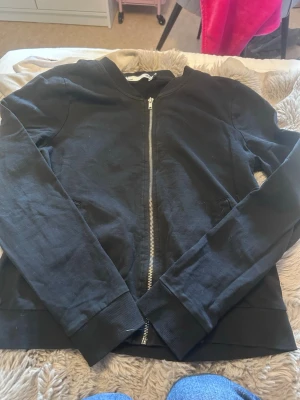 Svart hoodie med dragkedja - En enkel svart hoodie med dragkedja framtill och huva. Tillverkad i mjukt bomullsliknande material och har ribbade muddar vid ärmslut och nederkant. Perfekt för en avslappnad och stilren look.