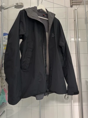 Svart vindjacka från Haglöfs - Snygg svart vindjacka från Haglöfs med dragkedja framtill och två sidofickor. Jackan har en hög krage och justerbar huva. Tillverkad i ett lätt och slitstarkt material som står emot vind och lätt regn. Perfekt för utomhusaktiviteter.