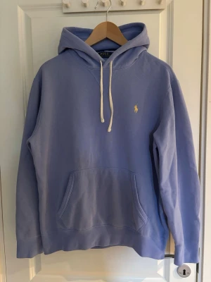 Ljusblå hoodie från Polo Ralph Lauren - Snygg ljusblå hoodie från Polo Ralph Lauren med klassisk huva och vita dragsnören. Tröjan har en känguruficka framtill och den ikoniska gula Polo-loggan broderad på bröstet. Tillverkad i mjuk bomull för extra komfort.