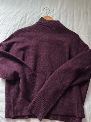 Vinröd stickad polotröja - Mysig vinröd stickad polotröja med lång ärm och ribbade muddar. Tröjan har en hög hals och rak passform, perfekt för kyliga dagar. Materialet ser ut att vara mjukt och lite luddigt, troligen ull- eller ullmix.