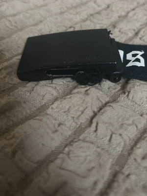 Svart bälte med Los Angeles-tryck - Snyggt svart bälte med vit text där det står 'Los Angeles' i gotisk stil. Bältet har ett brett band och ett svart metallspänne. Perfekt accessoar för att ge outfiten en streetwear-känsla.