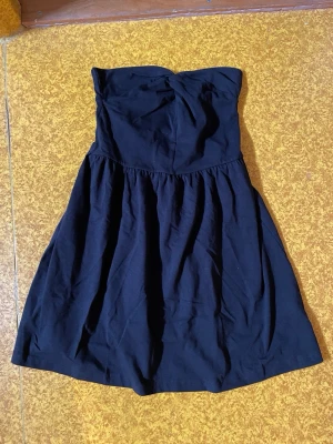 Svart axelbandslös kort klänning - Snygg svart axelbandslös klänning med utställd kjol och markerad midja. Klänningen har en enkel och stilren design som passar perfekt till många olika tillfällen. Tillverkad i mjukt tyg för en bekväm känsla.