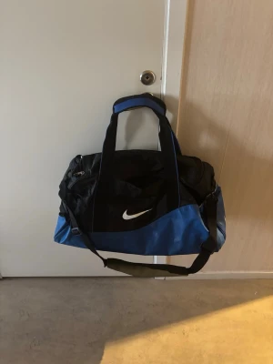 Blå och svart sportbag från Nike - Snygg sportbag från Nike i blått och svart med vit logga. Väskan har både bärhandtag och justerbar axelrem, samt flera fack med dragkedja. Perfekt för gymmet eller träningen när du vill ha plats för allt du behöver.