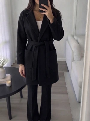 Svart kappa - Svart kappa med klassisk krage, långa ärmar och två stora fickor framtill. Modellen är rak med bälte. 