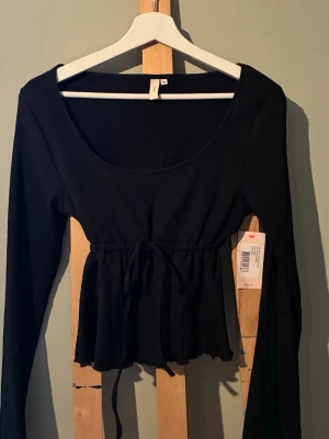 Svart långärmad topp från Nelly - Trendig svart långärmad topp från Nelly med rundad urringning och knyt i midjan. Toppen har en lätt peplum-effekt nedtill och är tillverkad i ett ribbat material som ger en snygg struktur. Priset går att prata om. Helt oanvänd då det inte var min stil men super gullig. 💕🖤