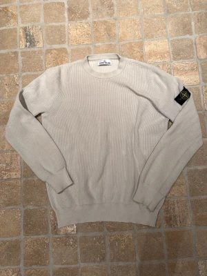 Tvär fet Stone Island tröja - Snygg beige stickad tröja från Stone Island med rund halsringning och ribbade muddar. Tröjan har den klassiska Stone Island-loggan på vänster ärm och är tillverkad i ett mjukt, strukturerat material. Perfekt för dig som gillar stilrena och tidlösa plagg.