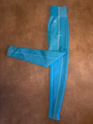 Turkosa seamless leggings strl S - Snygga turkosa  leggings med hög midja och vita detaljer längs benen. Stretchigt material som sitter tajt och bekvämt. Perfekta för träning eller chill. Gymshark logga på höften.