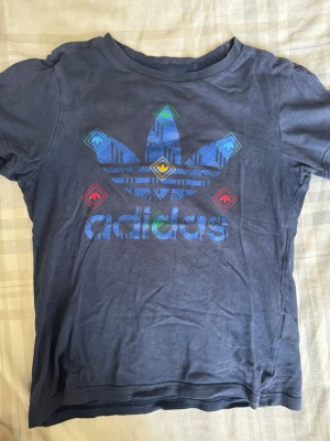 Blå Adidas baby tee med tryck - Säljer en blå t-shirt från Adidas med stort färgglatt tryck av Adidas-loggan på bröstet. T-shirten har korta ärmar och rund hals. Perfekt för dig som gillar sportig och avslappnad stil.