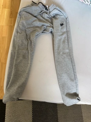 Gråa mjukisbyxor från Nike - Snygga gråa mjukisbyxor från Nike med svart logga på vänster lår. Byxorna har dragsko i midjan och muddar vid bensluten. Perfekta för chill dagar eller träning. Tillverkade i mjukt och bekvämt material.