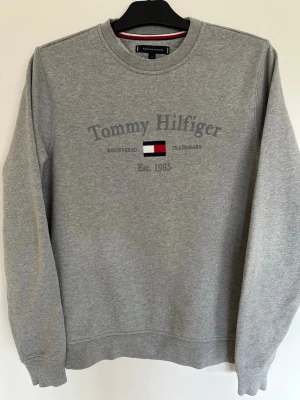 Grå sweatshirt Tommy Hilfiger - Ny/oanvänd klassisk grå sweatshirt från Tommy Hilfiger med rund halsringning och tryckt logga samt flagga på bröstet. Tröjan har långa ärmar och ribbade muddar vid ärmslut och nederkant. Perfekt för en avslappnad och stilren look. Liten broderad flagga även längst ner vid mudden på vänster arm. OBS! Prislapp/tagg SAKNAS! 