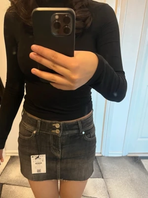 Mörkgrå kort jeanskjol med knappar - Snygg mörkgrå kort jeanskjol med dubbla knappar och klassiska fickor fram och bak. Kjolen har en rak passform och är tillverkad i ett robust denimtyg och har inbyggt shorts. Perfekt att matcha med en enkel topp för en trendig look.