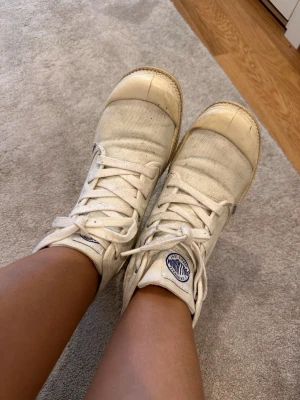 Vita Palladium high top sneakers - Säljer ett par vita high top sneakers från Palladium med klassisk logga på plösen. Skorna har en canvas-ovandel, vita snören och gummisula. Perfekta för dig som gillar en clean och avslappnad stil.
