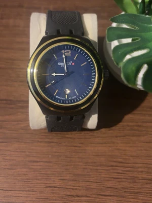 Klocka  - Stilren klocka med svart armband i gummi och blå urtavla. Klockan har gulddetaljer runt boetten, vita visare och datumvisning. Perfekt accessoar för dig som gillar en sportig och modern look. Priset kan diskuteras!