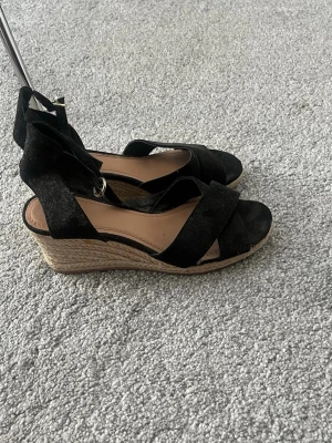 Svarta kilklackade sandaler med snörning - Säljer ett par svarta sandaler med kilklack och snörning runt ankeln. Ovandelen är i mockaimitation och sulan har flätad jutedesign som ger en somrig känsla. 