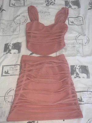 Rosa mesh set med topp och kjol - Säljer ett snyggt tvådelat set i rosa mesh. Setet består av en croppad topp med breda axelband och en matchande kjol med rynkade detaljer. Materialet är stretchigt och figurnära, perfekt för dig som gillar trendiga och feminina plagg.