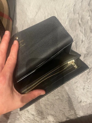 Svart plånbok i präglat läder - Snygg svart plånbok i präglat läder med klassiskt monogrammönster. Plånboken har guldfärgade detaljer, dragkedjefack och flera kortfack. Perfekt storlek för att få plats med allt du behöver och ändå hålla det stilrent.
