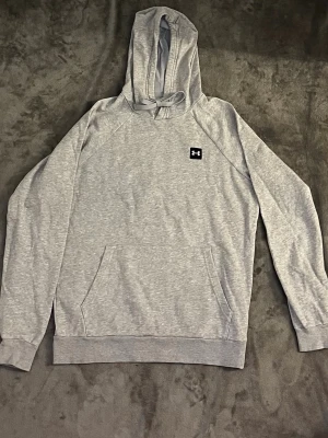 Grå hoodie från Under Armour - Snygg ljusgrå hoodie från Under Armour med klassisk huva och magficka. Diskret logga på bröstet och ribbade muddar vid ärmar och nederkant. Tillverkad i mjukt material som känns skönt mot huden. Perfekt för en avslappnad och sportig stil.