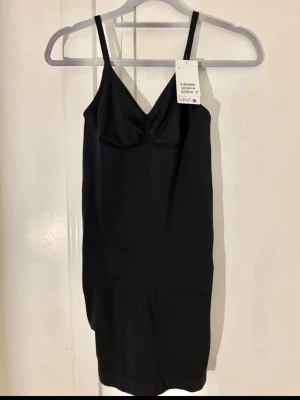 Svart shaping bodyklänning H&M - Säljer en svart shaping bodyklänning från H&M med smala, justerbara axelband och v-ringad urringning. Materialet är stretchigt och slätt, perfekt för att ge en smickrande siluett under kläder. Enkel och stilren design utan synliga sömmar.
