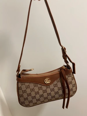  brun monogram axelremsväska - Säljer en snygg axelremsväska  i beige och brunt med klassiskt GG-monogram. Väskan har guldfärgad logga framtill, justerbar rem och detaljer i brunt läder. Perfekt storlek för det viktigaste och stängs med dragkedja. En riktig it-väska för dig som gillar lyxig stil.