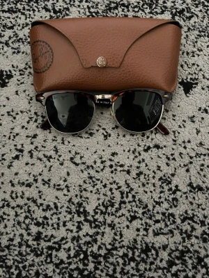 Ray-Ban Clubmaster solglasögon - Säljer ett par klassiska Ray-Ban Clubmaster solglasögon med svart ovandel och silvriga detaljer. Glasen är mörka och bågarna har en retroinspirerad form. Perfekt accessoar för soliga dagar!