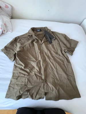 Beige kortärmad skjorta från Revolution - Snygg beige skjorta från Revolution med korta ärmar och klassisk krage. Skjortan har knappar framtill och två bröstfickor, varav ena är dekorerad med Revolution-loggan. Tillverkad i mjuk bomull, perfekt för varma dagar.