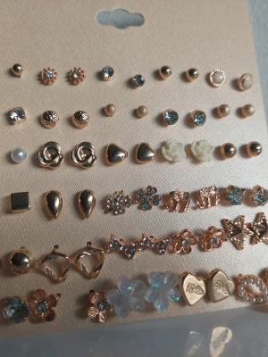 Set med 30 par örhängen i guldton - Säljer ett set med 30 par örhängen i olika former och stilar. Örhängena är i guldton med detaljer som blommor, hjärtan, stjärnor, pärlor och gnistrande stenar. Materialet är metall, plast och akryl. Perfekt för dig som vill variera din look med söta och trendiga detaljer.