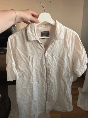 Beige linneskjorta med kort ärm - Säljer en beige skjorta i linne från Primark med klassisk krage och korta ärmar. Skjortan har knappar framtill och en avslappnad passform, perfekt för varma dagar.