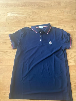 Marinblå pikétröja från Moncler - Snygg marinblå pikétröja från Moncler med klassisk krage och knappar framtill. Tröjan har röda och vita detaljer på krage och ärmslut samt Moncler-logga på bröstet. Perfekt för en stilren och sportig look.