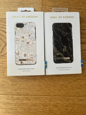 Två iDeal of Sweden mobilskal iPhone - Säljer två snygga mobilskal från iDeal of Sweden till iPhone SE/8/7/6/6S. Ett skal har ett elegant blommönster i vitt och beige (Pearl Blossom) och det andra har en stilren svart marmordesign med gulddetaljer (Golden Smoke Marble). Båda är tillverkade i hårdplast och kommer i originalförpackning.