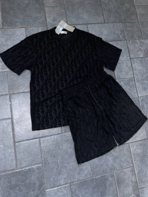 Svart set med mönster - Säljer ett svart set med t-shirt och shorts i mjukt material. Båda plaggen har ett diskret, upprepat mönster över hela ytan. T-shirten har rund hals och korta ärmar, shortsen har resår i midjan för skön passform. Perfekt för en avslappnad look, perfekt till sommaren.