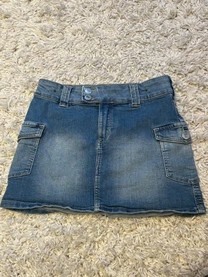 Jeanskjol - Snygg jeanskjol med låg midja. Kjolen har två knappar framtill och fina sidofickor med knappar. Obs är ganska liten i strl: midjemått rakt över 30 cm & kjol är ca 32 cm lång💙