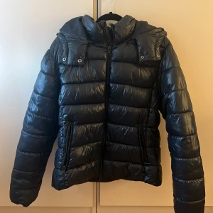 Mörkblå pufferjacka från 157 med huva - Säljer en mörkblå pufferjacka från 157 i storlek L. Jackan har en stor huva, dragkedja framtill och två fickor med dragkedja. Materialet är glansigt och känns riktigt mjukt. Perfekt för kalla dagar när du vill hålla dig varm och se snygg ut. Knappt använd.