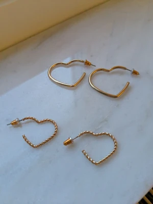 Två par hjärtformade örhängen i guld - Säljer två par örhängen i guldfärgad metall, båda i hjärtform. Ett par har en slät, enkel design medan det andra paret har en tvinnad struktur för en mer unik look. Perfekta för att mixa och matcha till olika outfits. Köpta i Munchen på en flygplats🌸