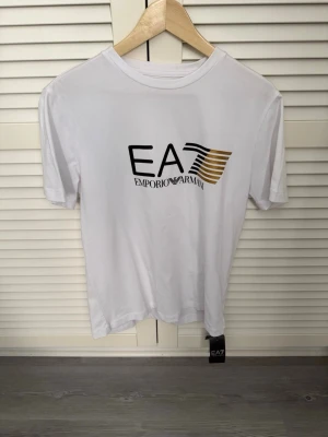 Vit EA7 Emporio Armani t-shirt helt ny - Snygg vit t-shirt från EA7 Emporio Armani med stor logga i svart och guld på bröstet. Klassisk rund hals och korta ärmar. Perfekt till jeans eller shorts för en clean och modern look. Helt ny Storelk L 