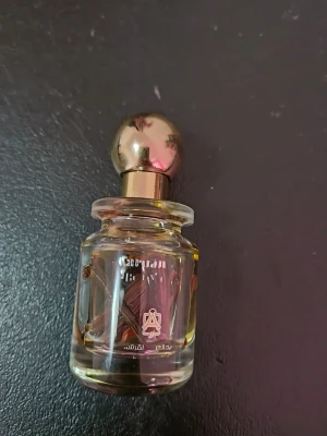 Exklusiv parfymflaska med gulddetaljer - Säljer en elegant parfym i en genomskinlig glasflaska med guldfärgat klotformat lock. Flaskan har dekorativa träbitar inuti och text i vitt samt arabiska tecken på framsidan. Perfekt för dig som gillar unika dofter och snygg design.