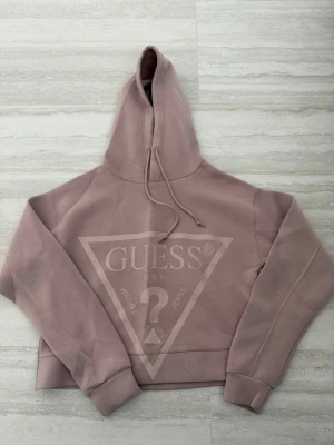 Ljusrosa hoodie från Guess - Säljer en ljusrosa hoodie från Guess med stor logga framtill. Hoodien har huva med dragsko och ribbade muddar vid ärmar och nederkant. Tillverkad i mjukt bomullsmaterial för en skön och avslappnad look.