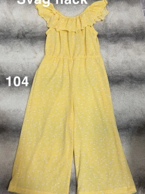Gul blommig jumpsuit med volang - Söt gul jumpsuit med små vita blommor och volangdetalj vid axlarna. Vida ben och resår i midjan för skön passform. Perfekt för dig som gillar en färgglad och somrig stil.🌸Använd en gång. Svag fläck. Storlek 104