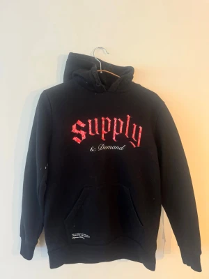 Svart hoodie Supply & Demand - Svart hoodie med huva och känguruficka från Supply & Demand. Stor röd text 'Supply' och vit text '& Demand' på bröstet. Klassisk passform med ribbade muddar och mjukt material, perfekt för en avslappnad streetwear-look.