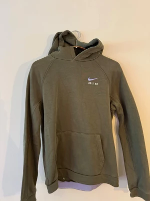 Olivgrön Nike Air hoodie med huva - Säljer en olivgrön Nike Air hoodie med stor ficka framtill och klassisk huva. Nike-logga och AIR-tryck både på bröstet och ryggen. Hoodien har långa ärmar och är gjord i mjukt material, perfekt för en avslappnad streetwear-look.